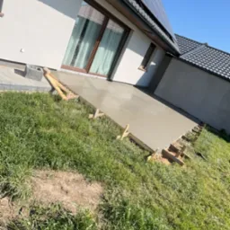 Wylany fundament pod taras domu jednorodzinnego z panelami słonecznymi na dachu, widoczne drewniane szalunki i trawa.