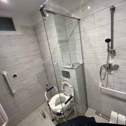 Dastin Burzyński - Nowoczesna łazienka z kabiną prysznicową i toaletą podwyższoną z poręczami, przystosowana dla osób z ograniczeniami ruchowymi. Widoczne białe płytki imitujące cegłę oraz chromowana armatura.