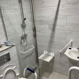 Dastin Burzyński - Łazienka z udogodnieniami dla osób niepełnosprawnych: prysznic z siedziskiem, umywalka z uchwytami, toaleta z poręczą, wykończona płytkami imitującymi drewno.