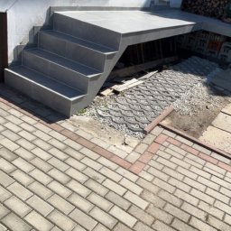 Dastin Burzyński - Nowoczesne, szare schody zewnętrzne z płytek prowadzące do budynku z widocznym oknem i stertą drewna w tle. Częściowo ułożona kostka brukowa na podłożu.