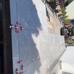 Dastin Burzyński - Układanie szarych płytek na tarasie z użyciem czerwonych i białych krzyżyków dystansowych, widoczny fragment ogrodzenia i osoba w tle.