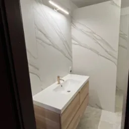 Nowoczesna łazienka z podświetlanym lustrem, umywalką nablatową z drewnianą szafką z szufladami i marmurowymi płytkami na ścianach, widziana zza framugi drzwi.