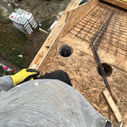 Dastin Burzyński - Z góry widok na deskowanie zbrojone siatką stalową, przygotowane pod wylewkę, z widocznymi otworami na słupy, w tle pracownik miesza beton w wiadrach.