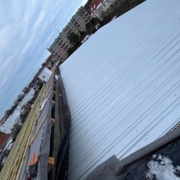 Dastin Burzyński - Montaż białego, blaszanej pokrycia dachowego z widoczną konstrukcją drewnianą i membraną w tle zabudowa miejska.