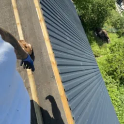 Montaż blachy trapezowej na dachu z widocznymi łatami drewnianymi, ręką w niebieskiej rękawicy trzymającą brązową paczkę, cień osoby na dachu.