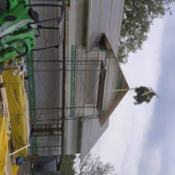 Budowa domu w stanie surowym otwartym z zawieszoną wiechą na szczycie dachu, widoczne rusztowania, elementy konstrukcyjne, deski i mała zielona koparka.