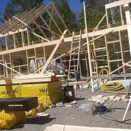 Dastin Burzyński - Wznoszenie szkieletu drewnianego domu z widocznymi belkami stropowymi, żółtymi izolacjami Valbo Tra i rozłożonymi narzędziami na żwirowym podłożu.