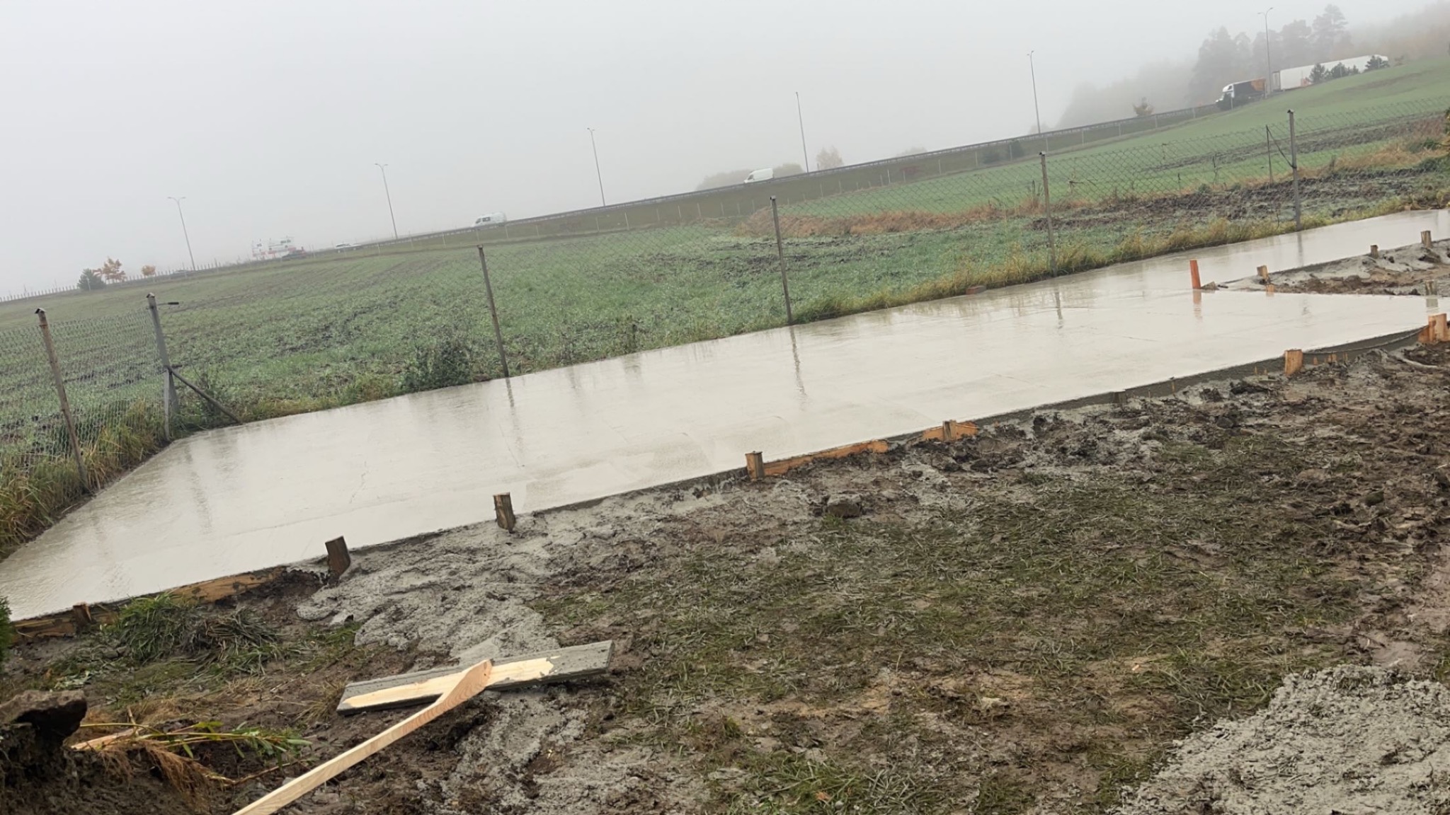 Świeżo wylana, mokra betonowa płyta fundamentowa z drewnianym szalunkiem, widoczna na tle pochmurnego krajobrazu z drogą szybkiego ruchu w oddali.