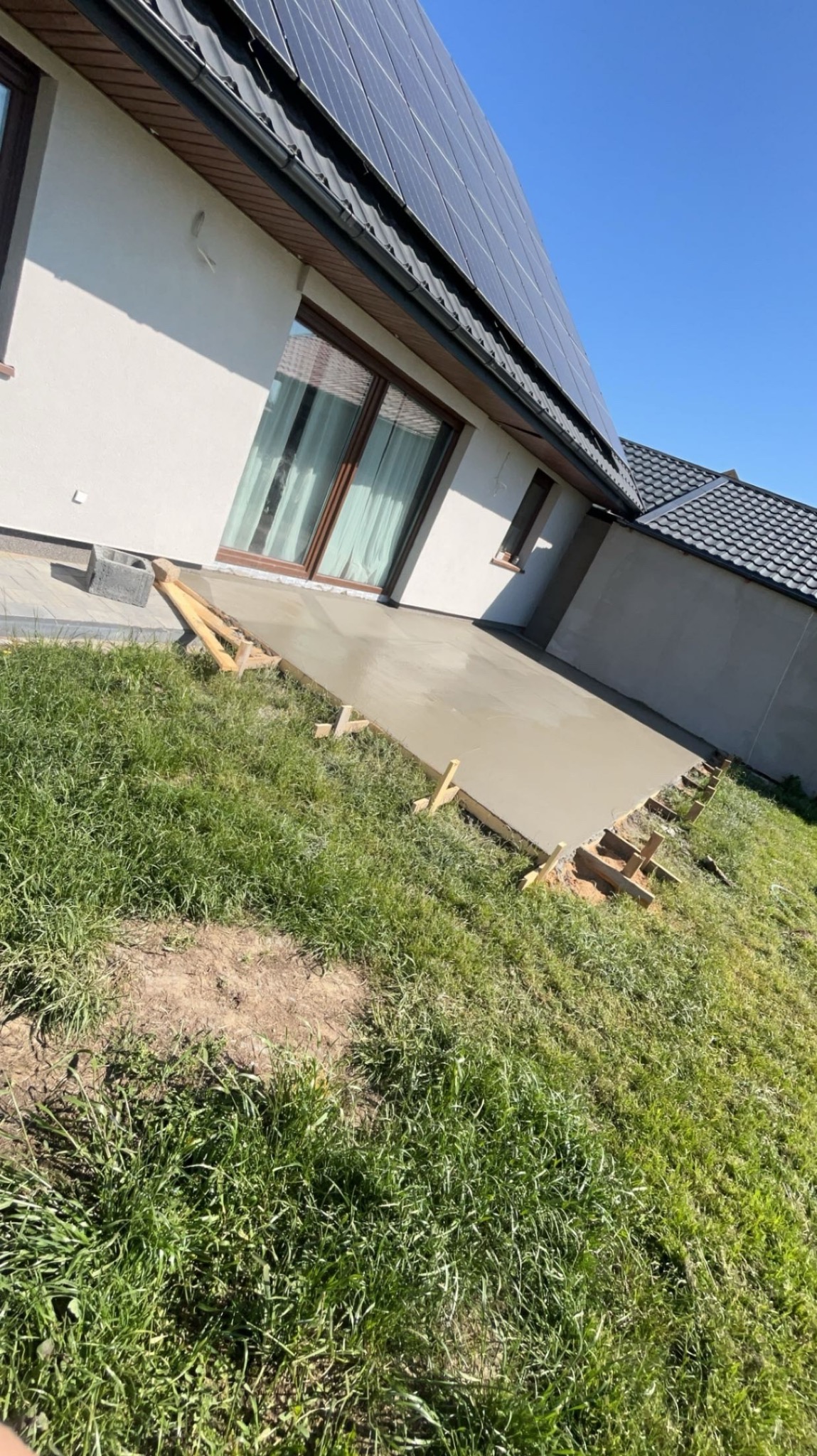 Wylany fundament pod taras domu jednorodzinnego z panelami słonecznymi na dachu, widoczne drewniane szalunki i trawa.