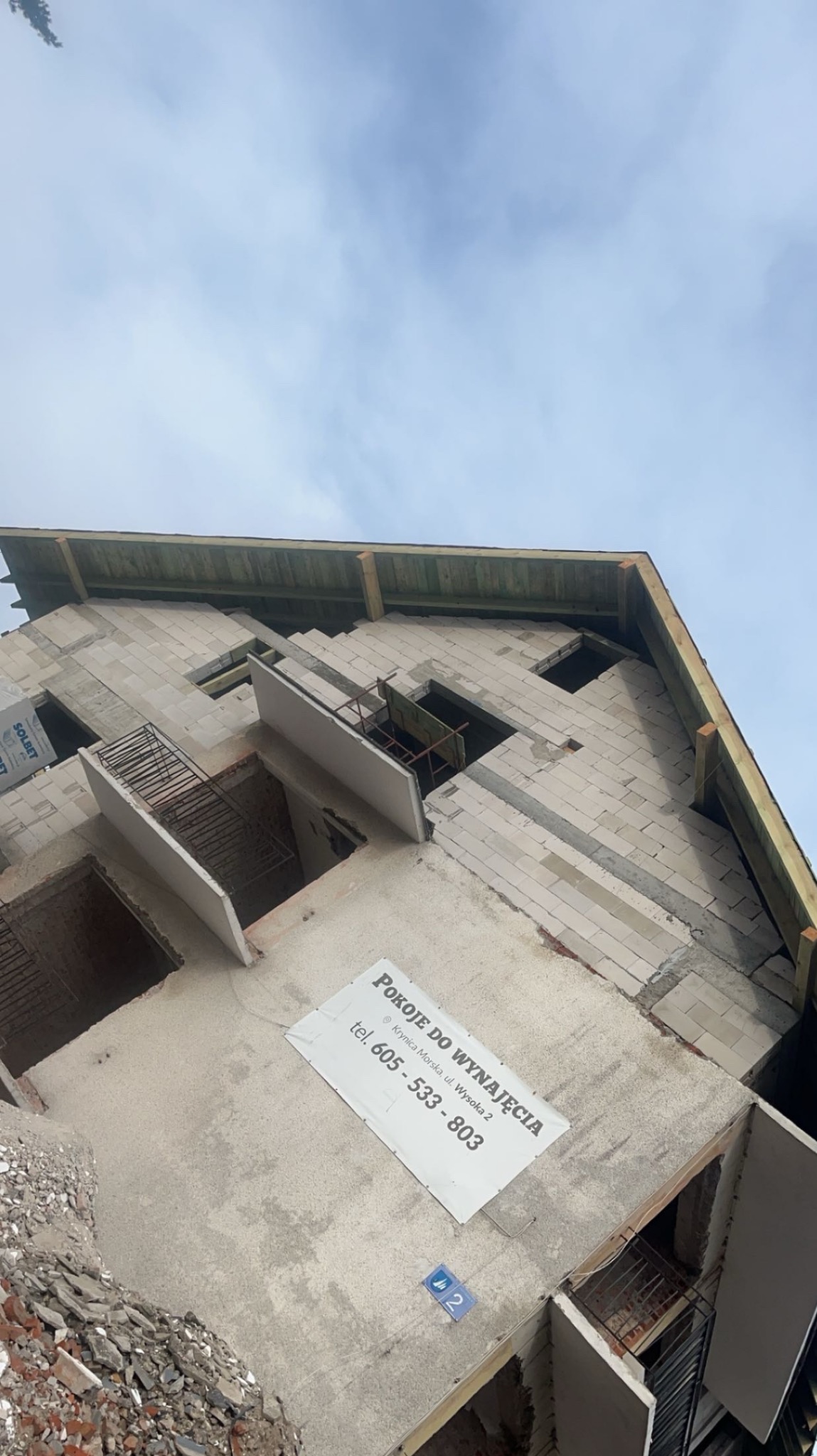 Niedokończony budynek z betonu komórkowego z widocznymi elementami konstrukcyjnymi, drewnianą więźbą dachową i banerem reklamowym 'Pokoje do wynajęcia' na ścianie.