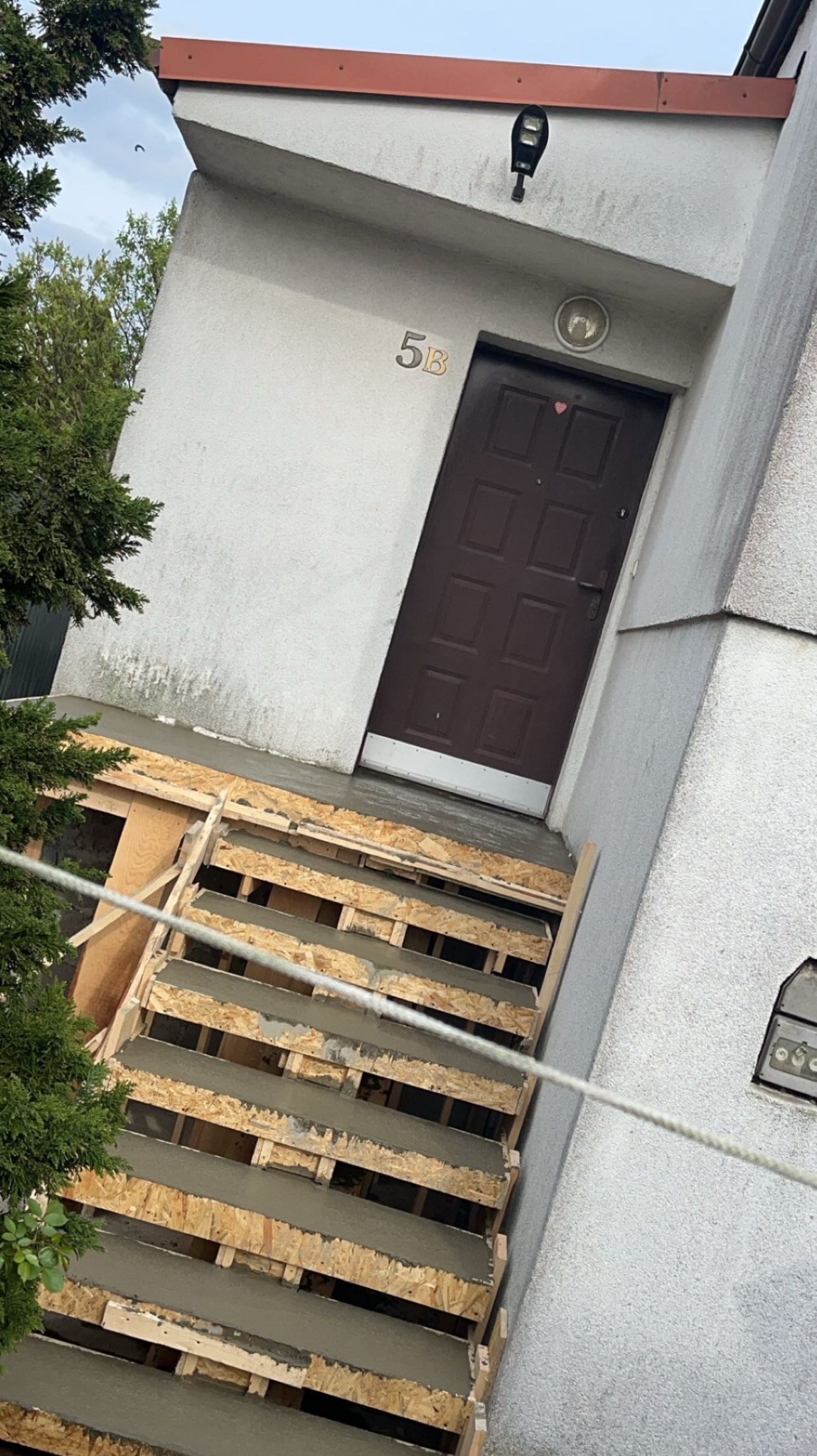 Budowa betonowych schodów wejściowych do domu z numerem 5B, widoczne szalunki wypełnione świeżym betonem, oświetlenie zewnętrzne nad drzwiami.