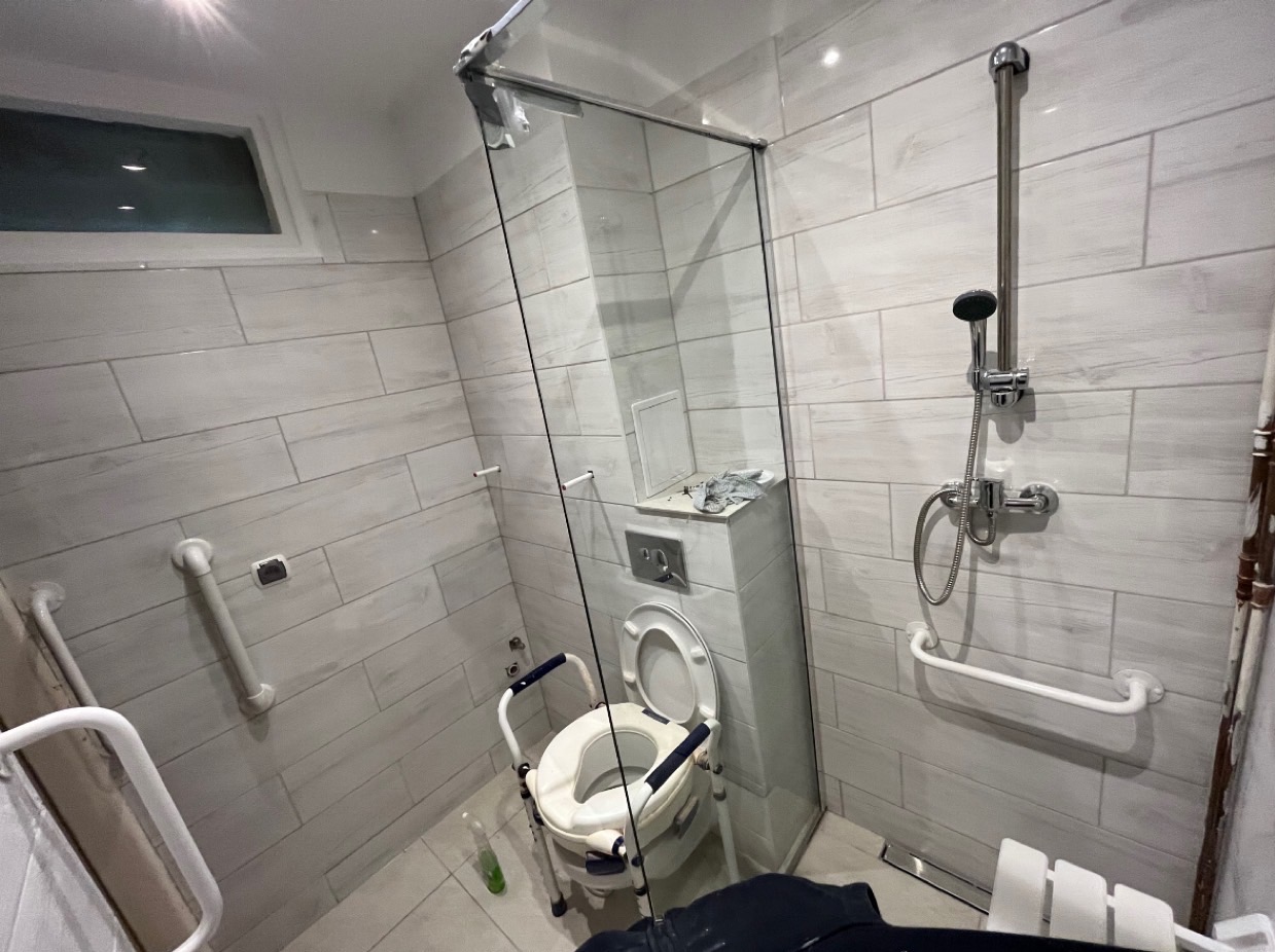 Nowoczesna łazienka z kabiną prysznicową i toaletą podwyższoną z poręczami, przystosowana dla osób z ograniczeniami ruchowymi. Widoczne białe płytki imitujące cegłę oraz chromowana armatura.