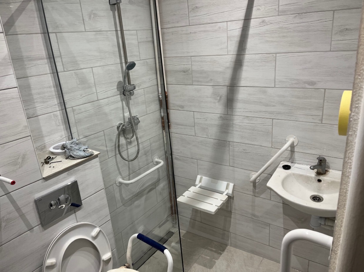 Łazienka z udogodnieniami dla osób niepełnosprawnych: prysznic z siedziskiem, umywalka z uchwytami, toaleta z poręczą, wykończona płytkami imitującymi drewno.