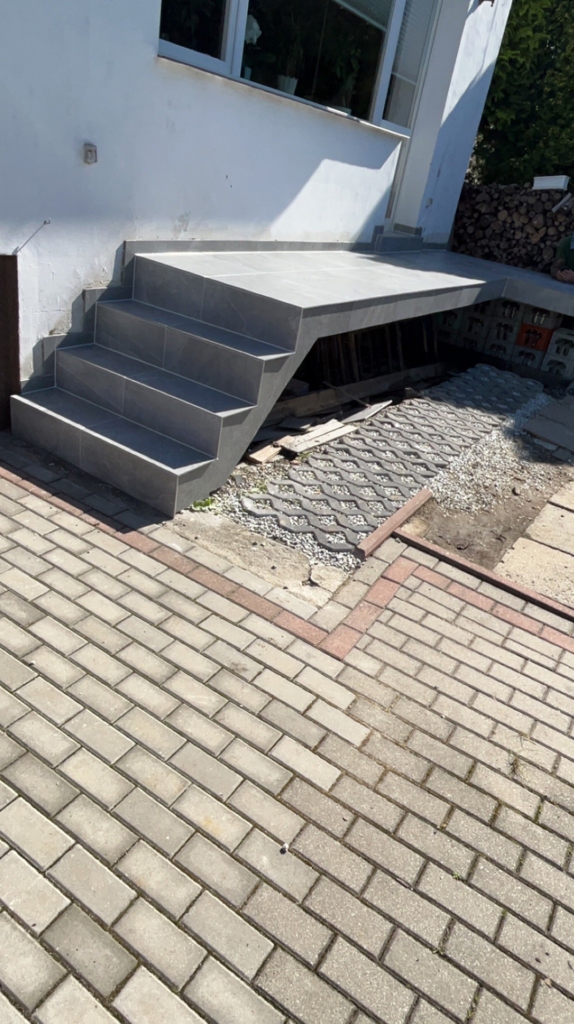 Nowoczesne, szare schody zewnętrzne z płytek prowadzące do budynku z widocznym oknem i stertą drewna w tle. Częściowo ułożona kostka brukowa na podłożu.