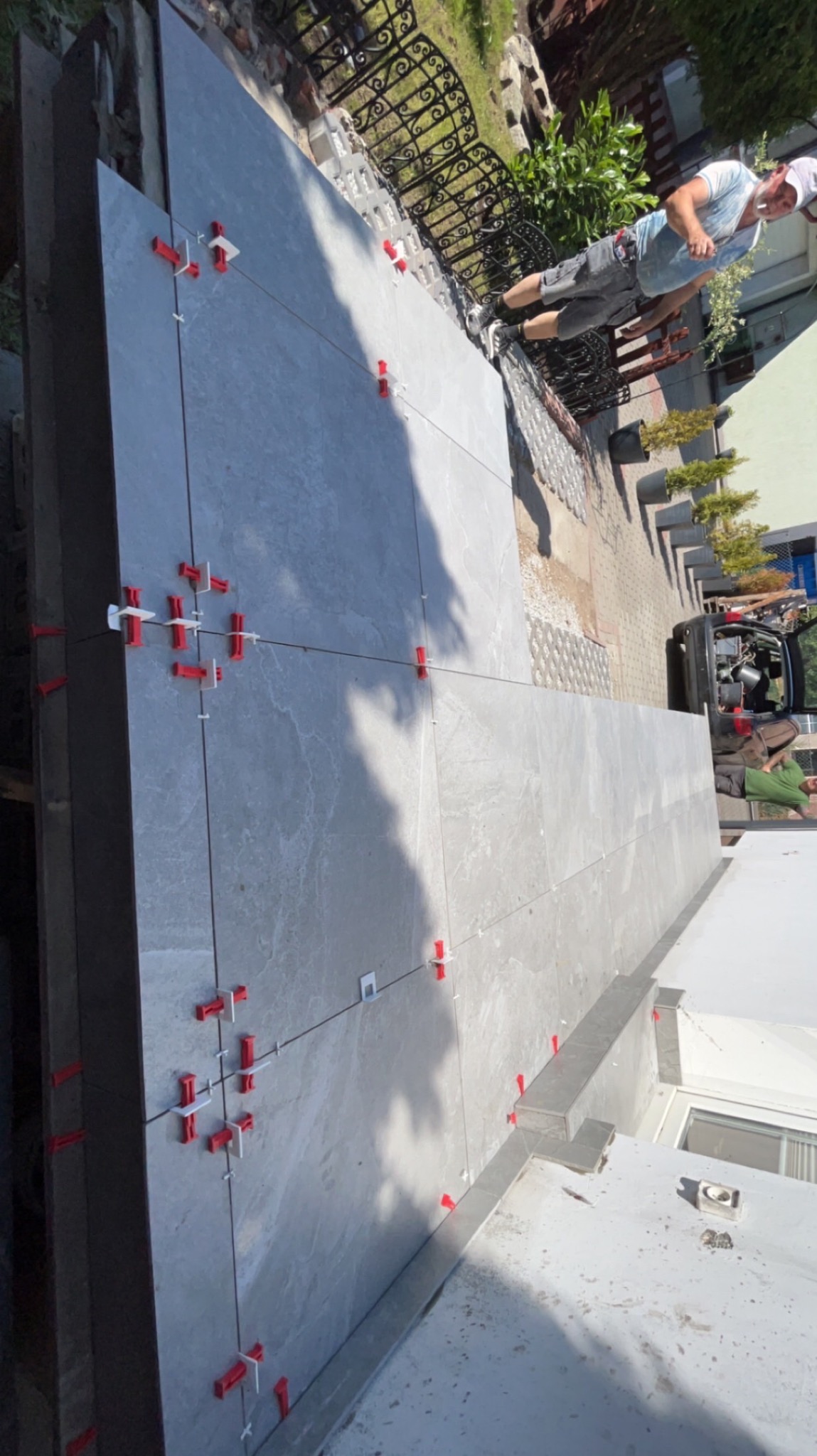 Układanie szarych płytek na tarasie z użyciem czerwonych i białych krzyżyków dystansowych, widoczny fragment ogrodzenia i osoba w tle.