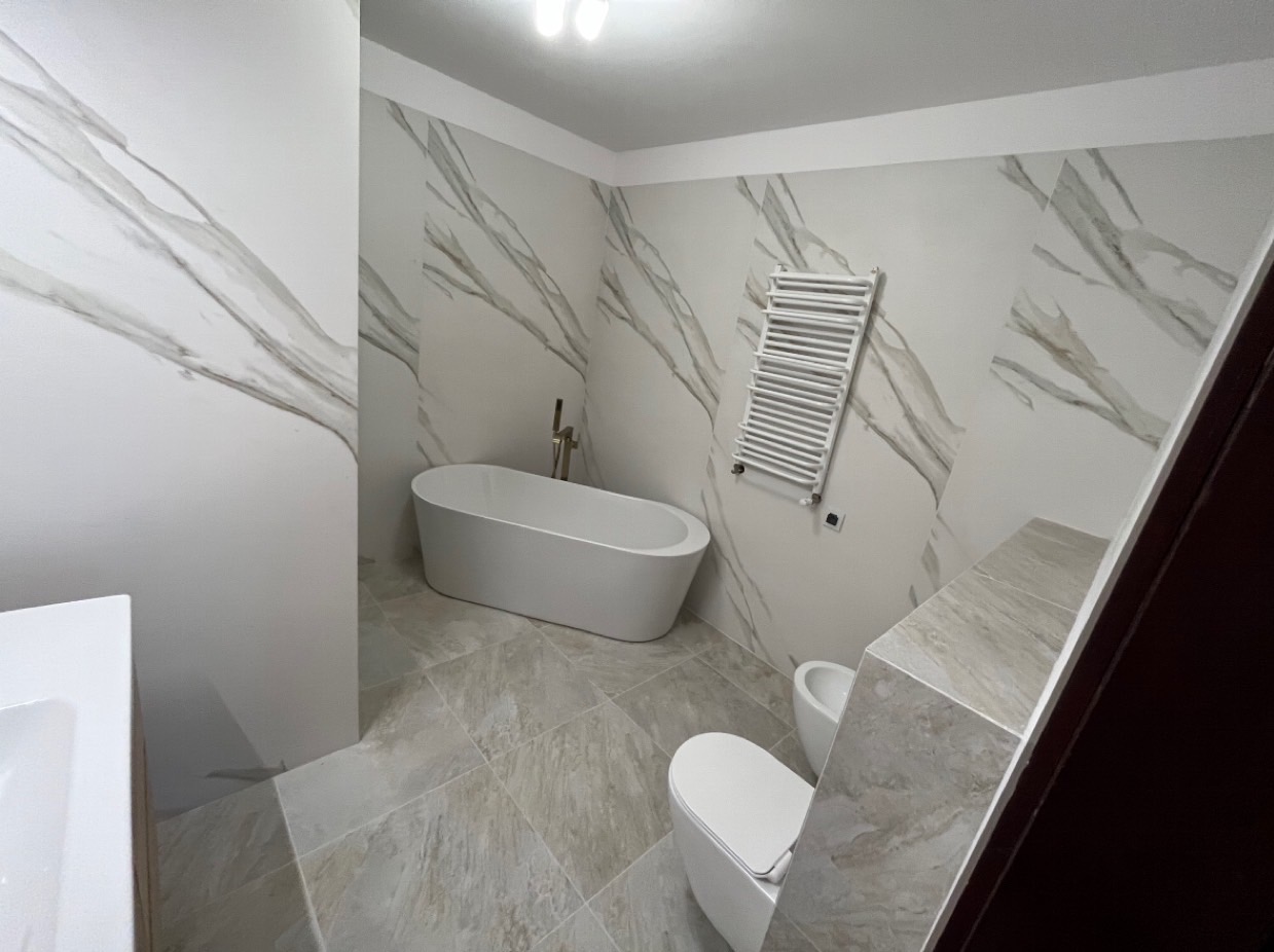 Nowoczesna łazienka z wolnostojącą białą wanną, białym grzejnikiem drabinkowym, bidetem i toaletą, wykończona płytkami imitującymi marmur z szarymi żyłami.