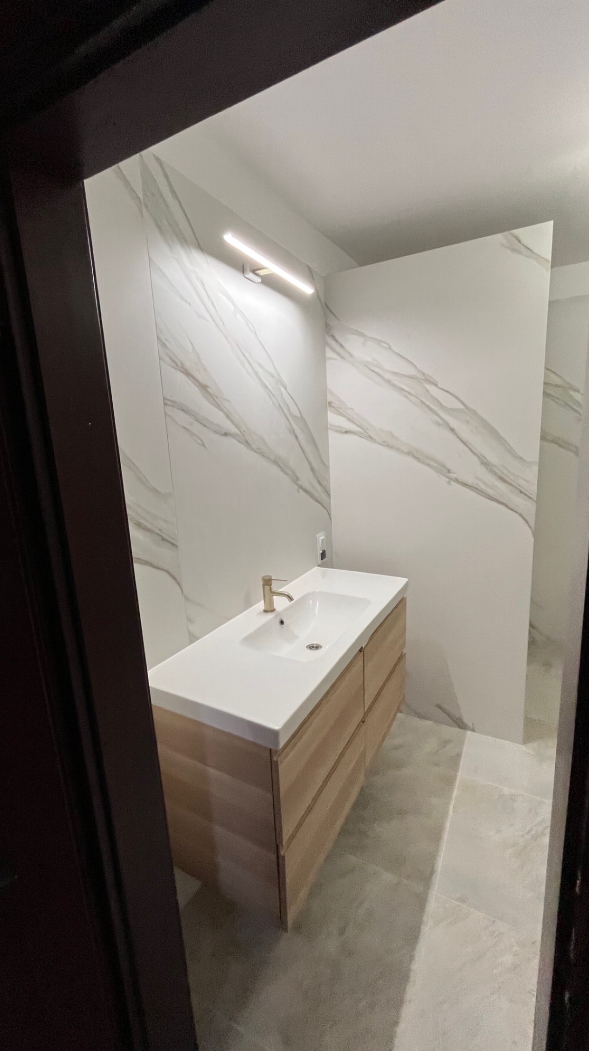 Nowoczesna łazienka z podświetlanym lustrem, umywalką nablatową z drewnianą szafką z szufladami i marmurowymi płytkami na ścianach, widziana zza framugi drzwi.