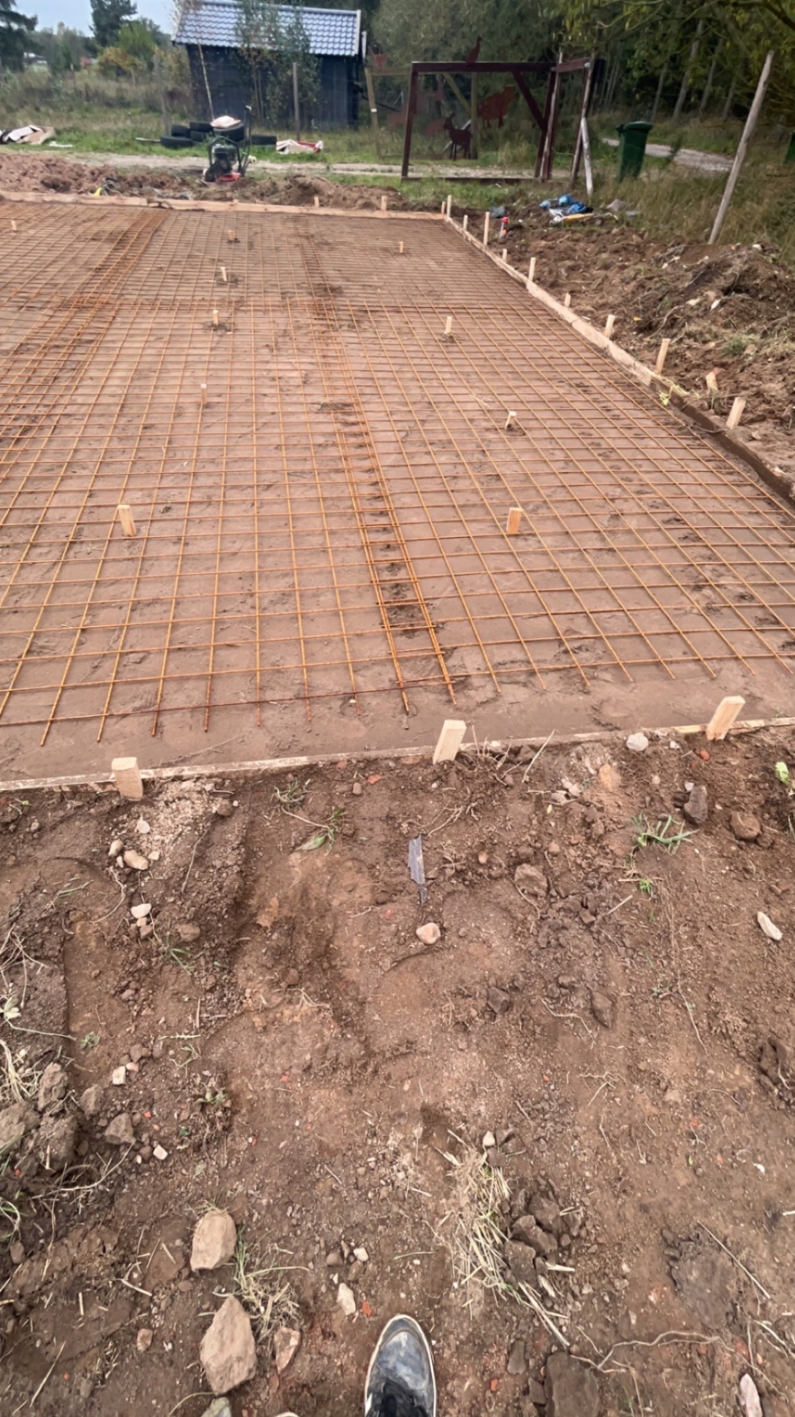 Zbrojenie fundamentu z siatki stalowej na przygotowanym podłożu, widoczne drewniane szalunki i poziomujące kołki, w tle zarys budynku gospodarczego.