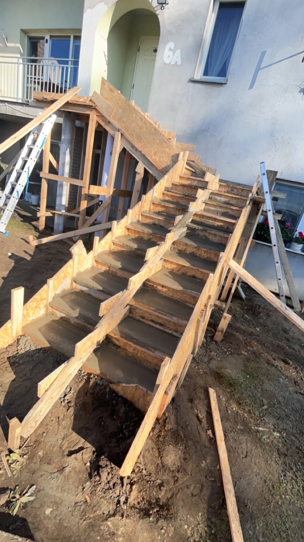 Budowa betonowych schodów zewnętrznych do domu, widoczne drewniane szalunki wypełnione świeżym betonem, drabiny opierają się o konstrukcję, ziemia wokół wykopana.