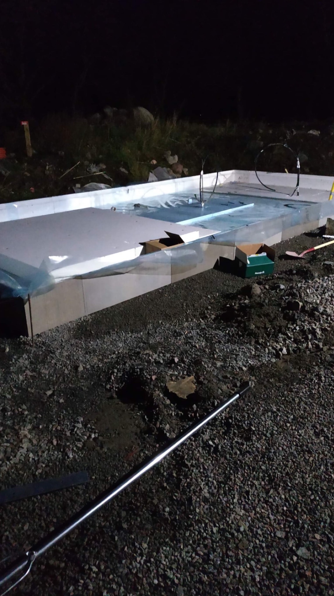 Przygotowany, izolowany fundament płyty na żwirze w ciemnych warunkach oświetleniowych, widoczne wystające rurki instalacji, kartony i narzędzia budowlane.