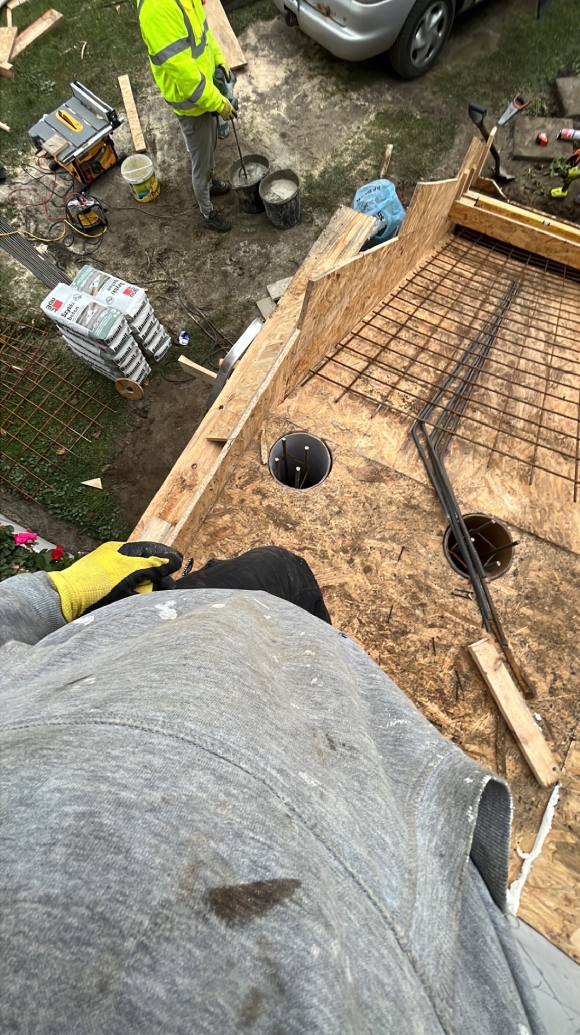 Z góry widok na deskowanie zbrojone siatką stalową, przygotowane pod wylewkę, z widocznymi otworami na słupy, w tle pracownik miesza beton w wiadrach.