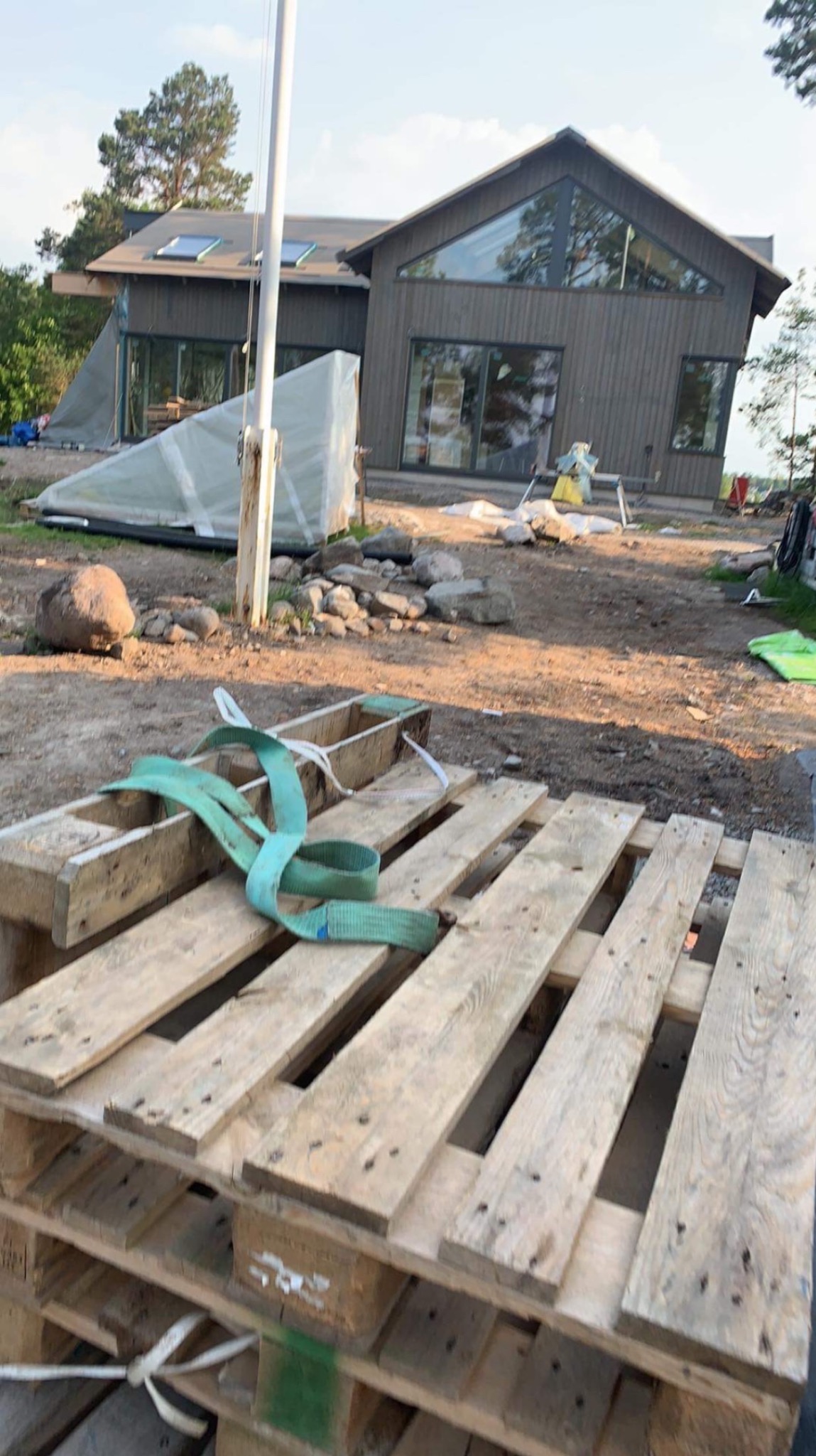 Nowoczesny dom z dużymi oknami i skośnym dachem w trakcie budowy, widoczne materiały budowlane i palety na pierwszym planie.