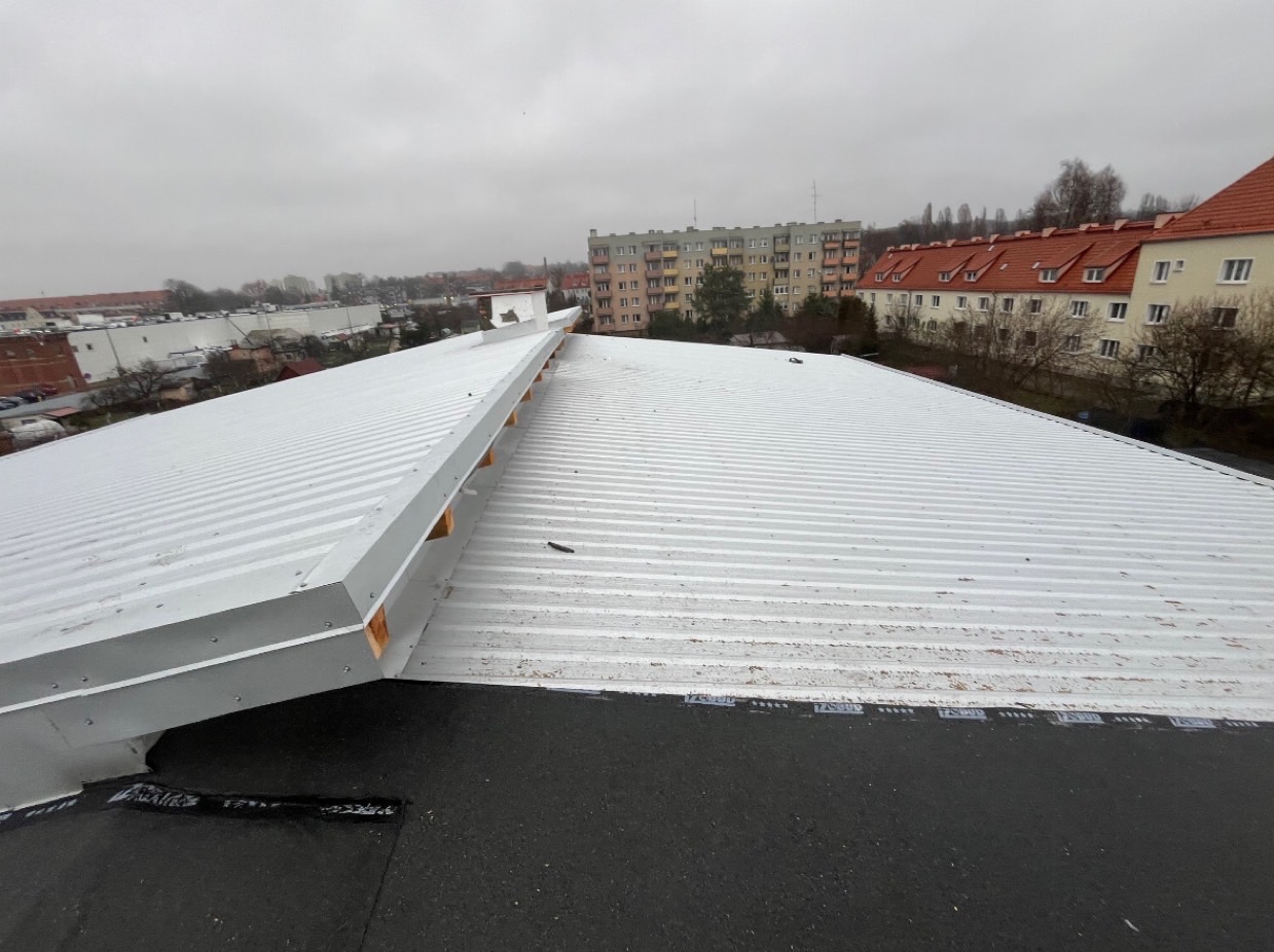 Widok z góry na duży, płaski dach pokryty blachą trapezową w kolorze białym, z widocznymi elementami konstrukcyjnymi i fragmentem papy na krawędzi, w tle budynki mieszkalne w pochmurny dzień.
