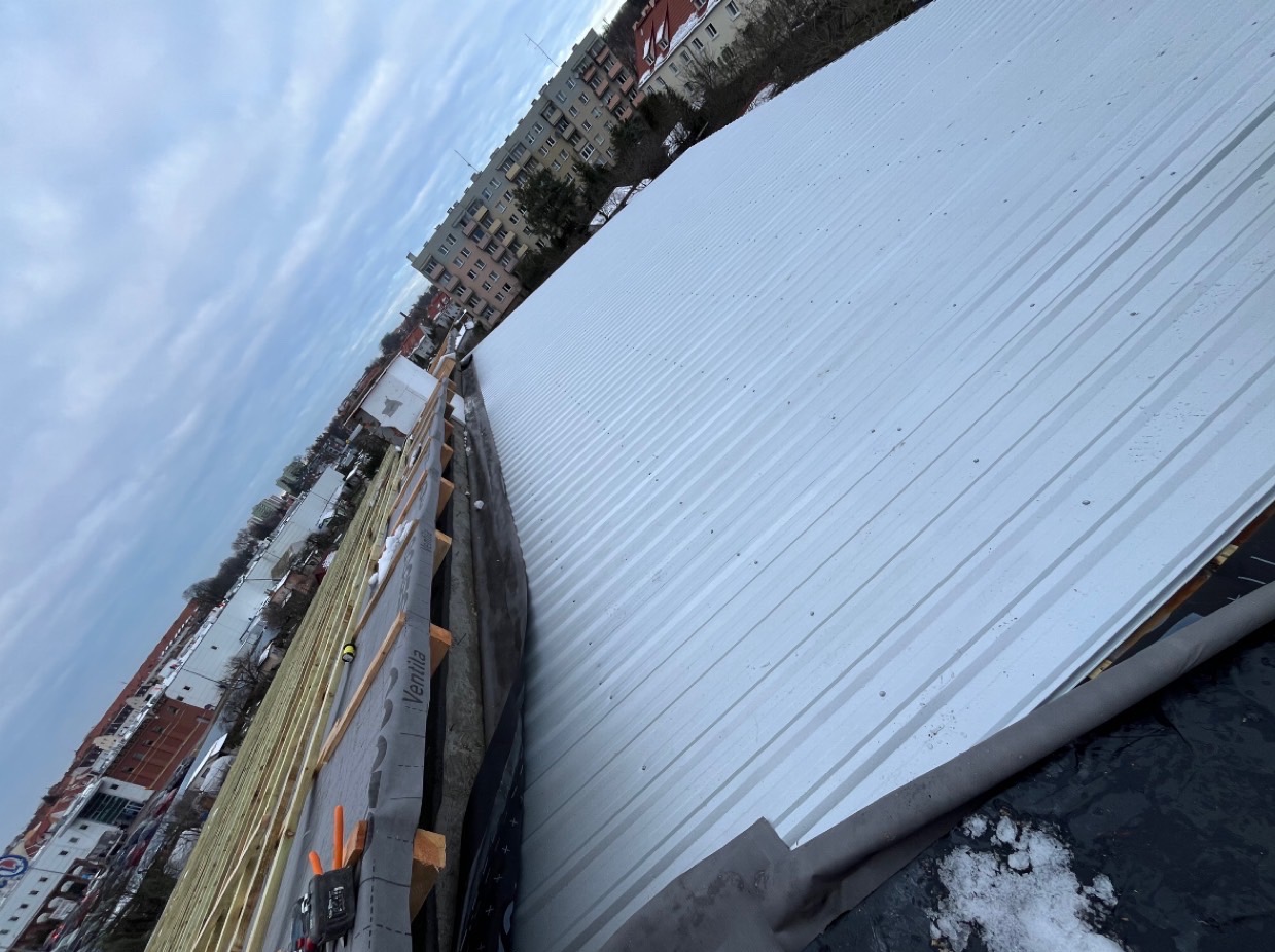 Montaż białego, blaszanej pokrycia dachowego z widoczną konstrukcją drewnianą i membraną w tle zabudowa miejska.