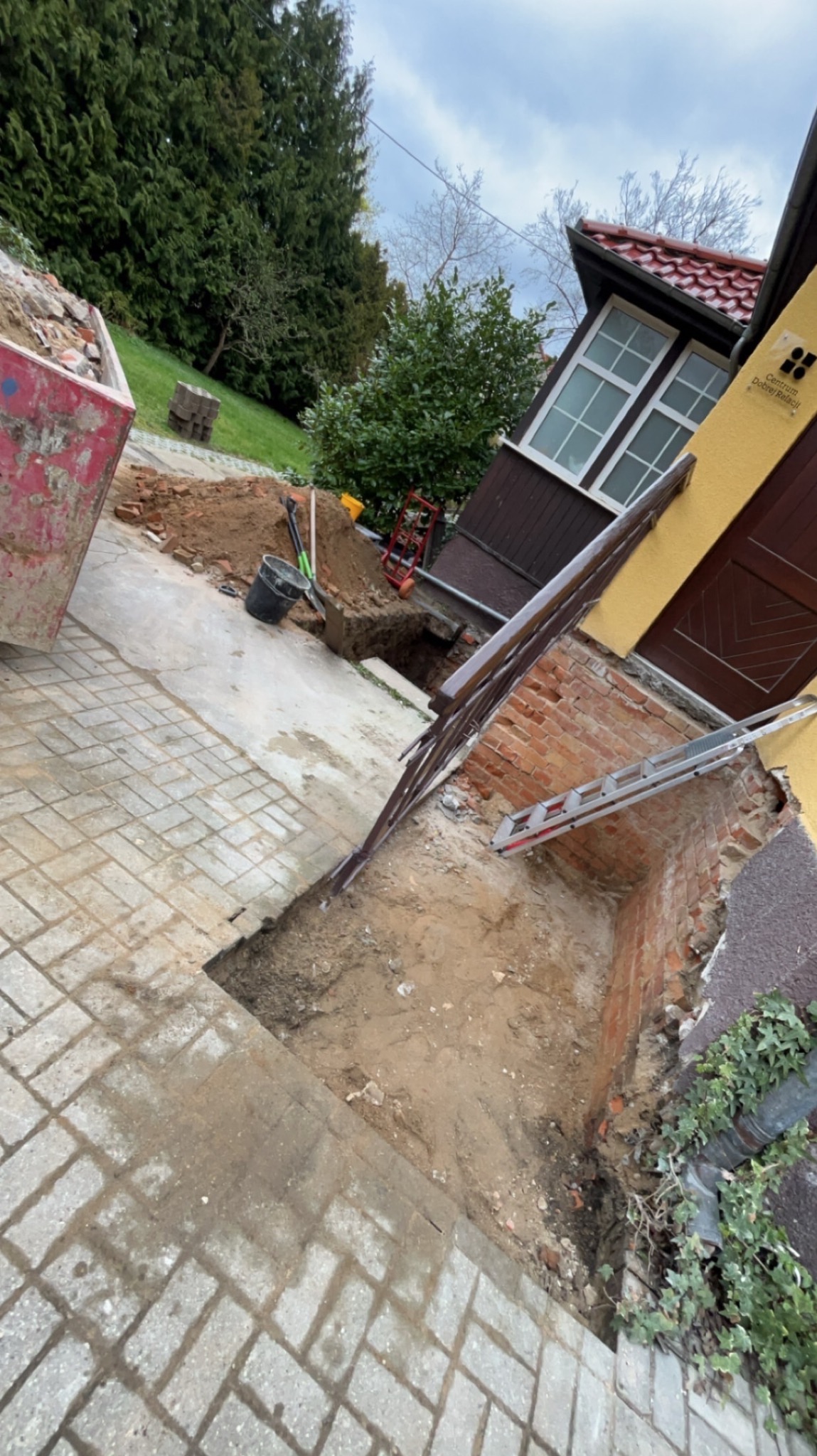 Wykop fundamentów przy ścianie budynku z cegły, widoczny kontener na gruz, drabina oparta o ścianę, narzędzia budowlane w wykopie.