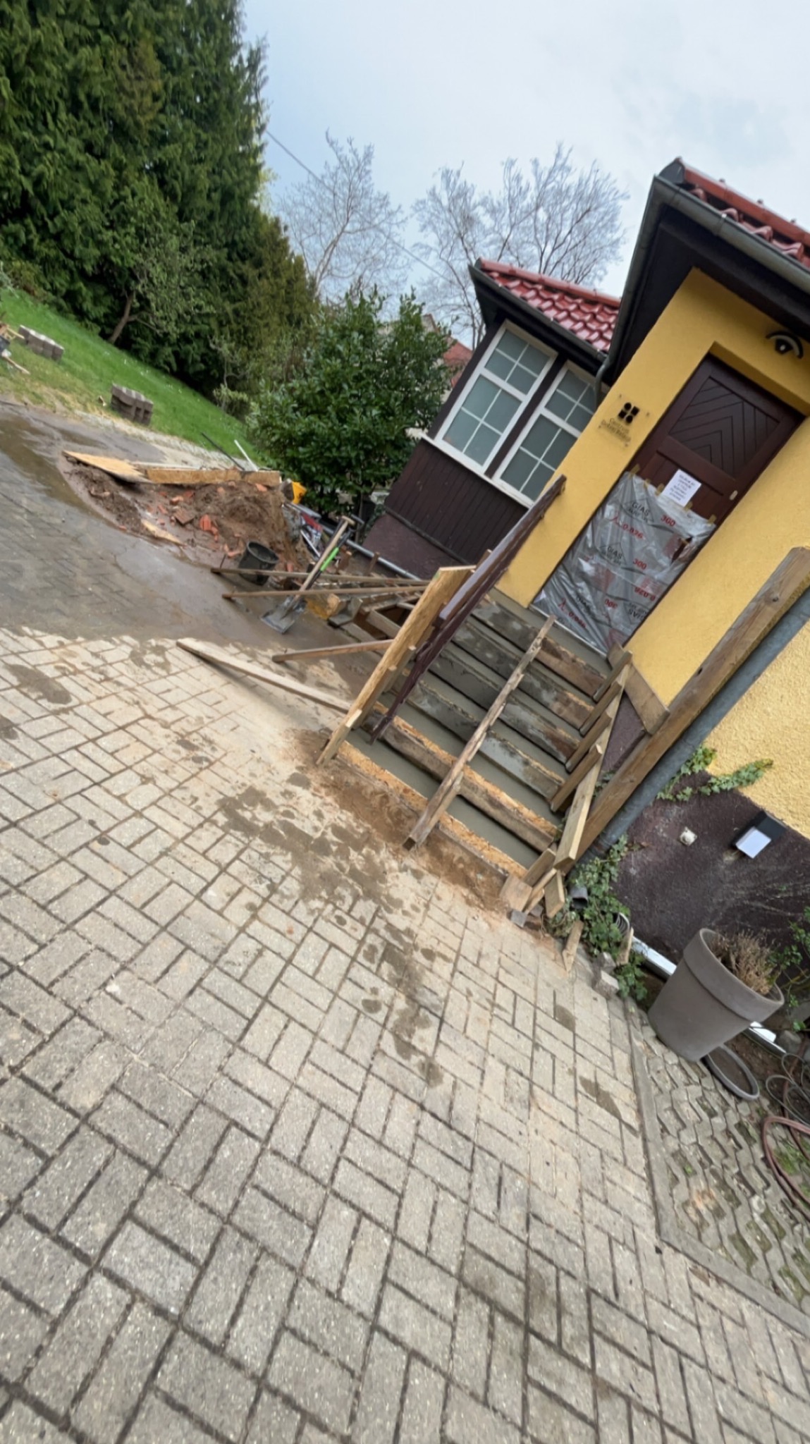 Budowa betonowych schodów wejściowych do żółtego domu z czerwoną dachówką, widoczne szalunki i mokry beton, teren budowy z narzędziami i materiałami.