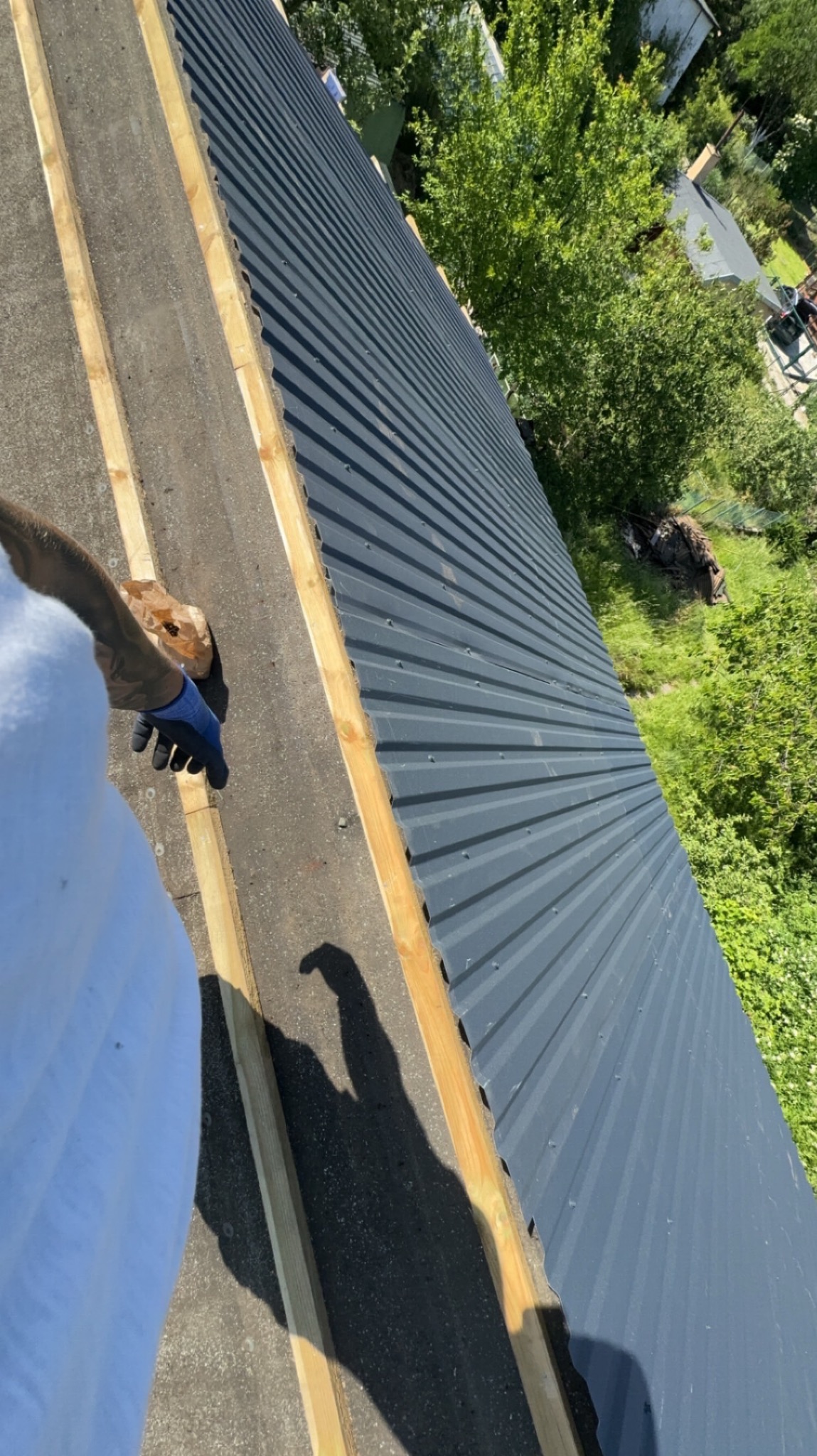 Montaż blachy trapezowej na dachu z widocznymi łatami drewnianymi, ręką w niebieskiej rękawicy trzymającą brązową paczkę, cień osoby na dachu.