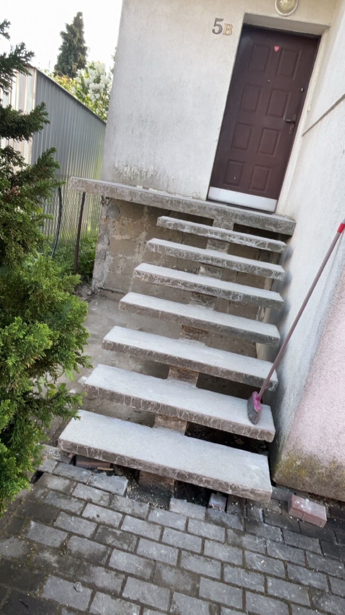 Betonowe schody wejściowe do budynku z numerem 5B, z widocznymi ubytkami w podłożu i szczotką opartą o ścianę, obok rośnie tuja.
