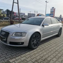 Srebrny sedan Audi A8 na kostce brukowej, czarne felgi, w tle budynki i reklamy zewnętrzne.