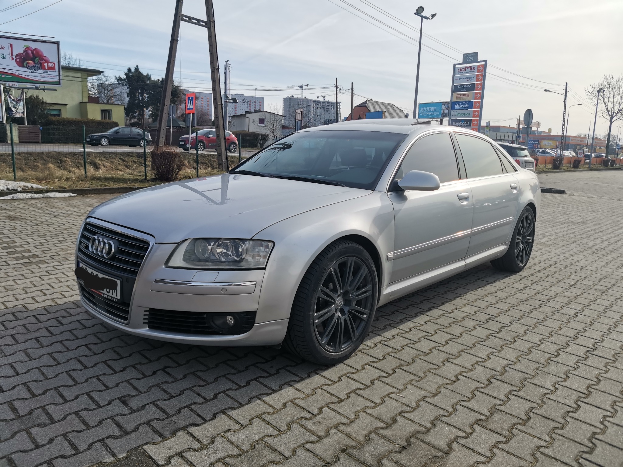 Srebrny sedan Audi A8 na kostce brukowej, czarne felgi, w tle budynki i reklamy zewnętrzne.