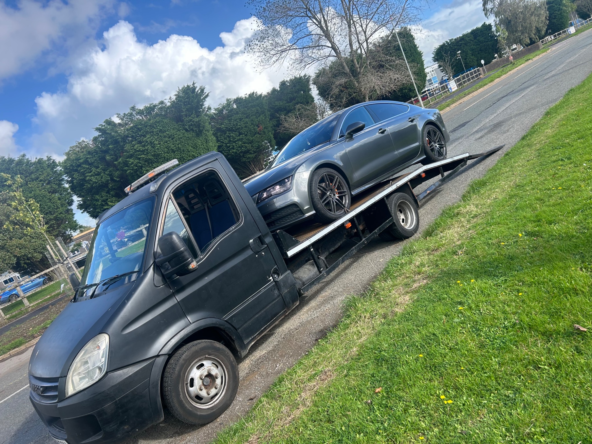 Czarna laweta z szarym samochodem osobowym typu sedan na platformie, zaparkowana na poboczu drogi z trawiastym terenem w tle, widok z boku.