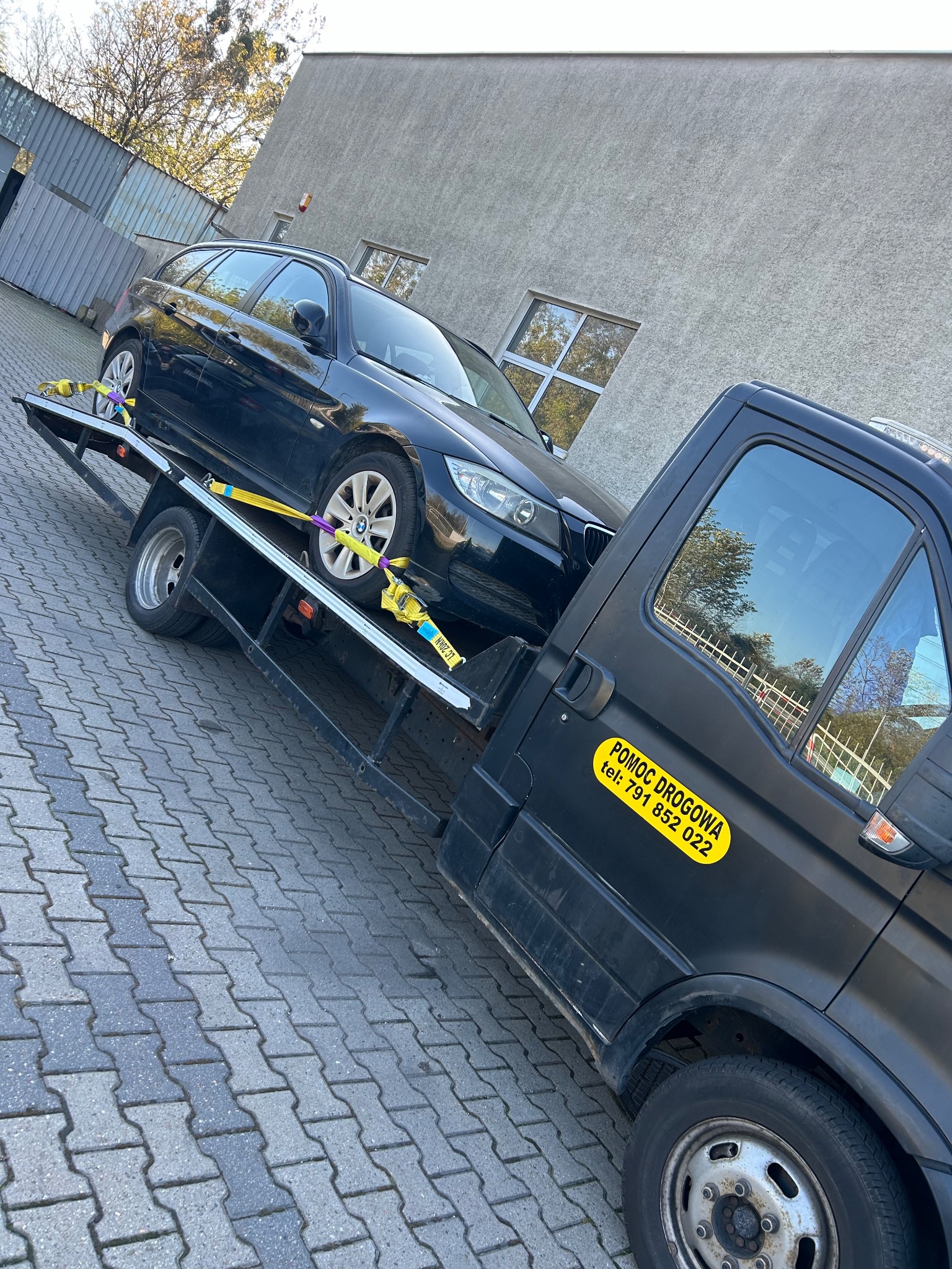 Czarny samochód osobowy zabezpieczony żółtymi pasami transportowymi na platformie pojazdu pomocy drogowej z naklejką informacyjną 'Pomoc Drogowa tel: 791 852 022'.