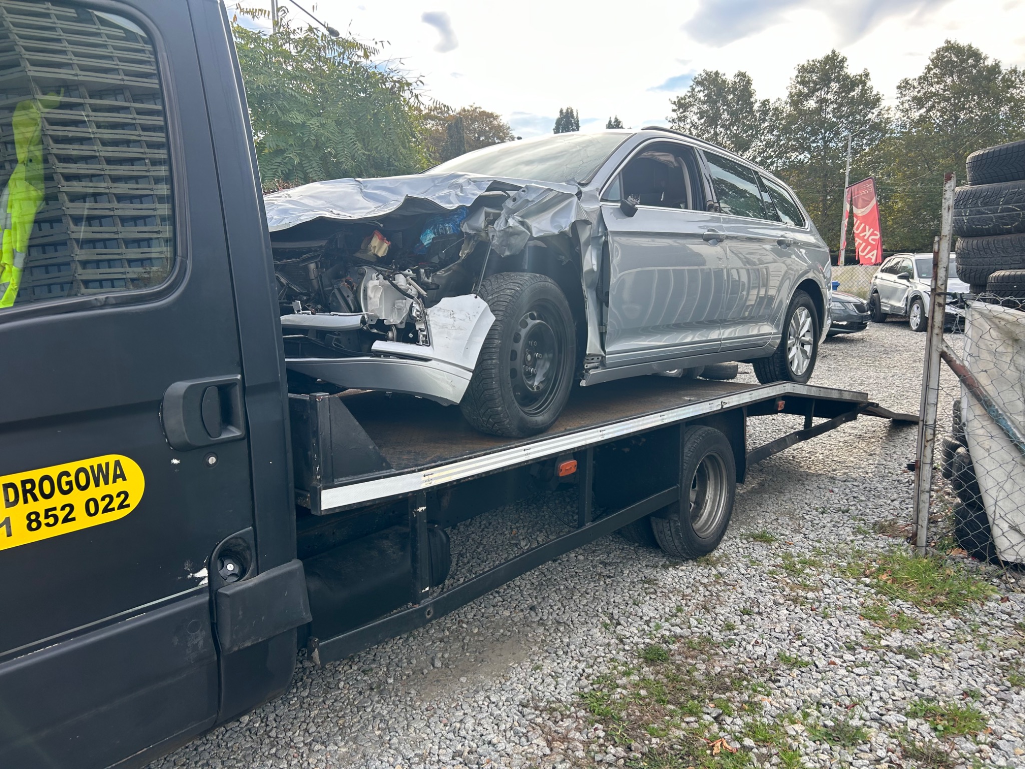 Srebrny samochód kombi z poważnymi uszkodzeniami przedniej części, załadowany na platformę pojazdu pomocy drogowej z żółtym napisem 'Pomoc Drogowa' i numerem telefonu, widok z boku na żwirowym...
