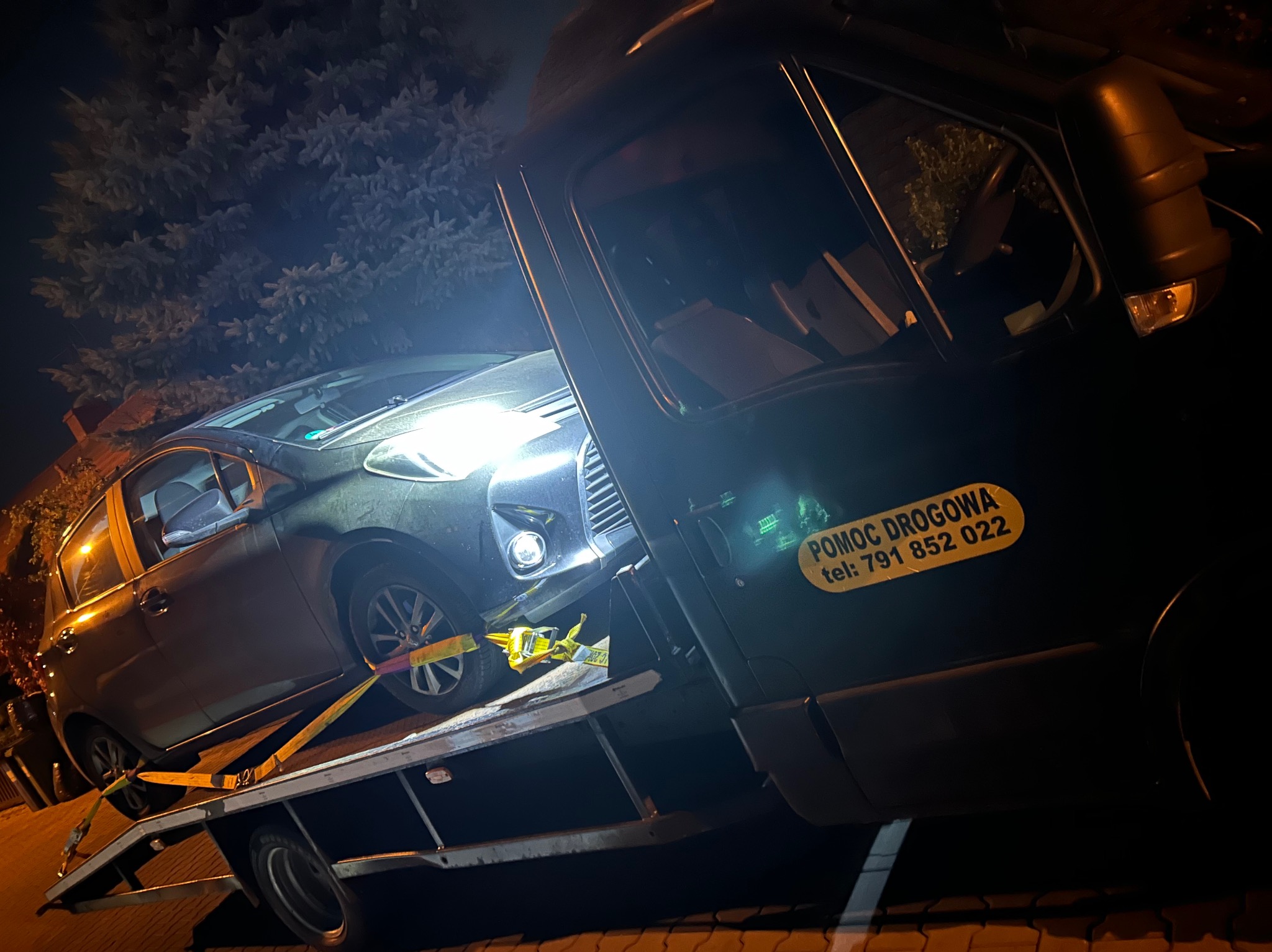 Samochód osobowy zabezpieczony pasami transportowymi na platformie pojazdu pomocy drogowej z naklejką informacyjną 'Pomoc Drogowa' i numerem telefonu, nocą na tle drzew.