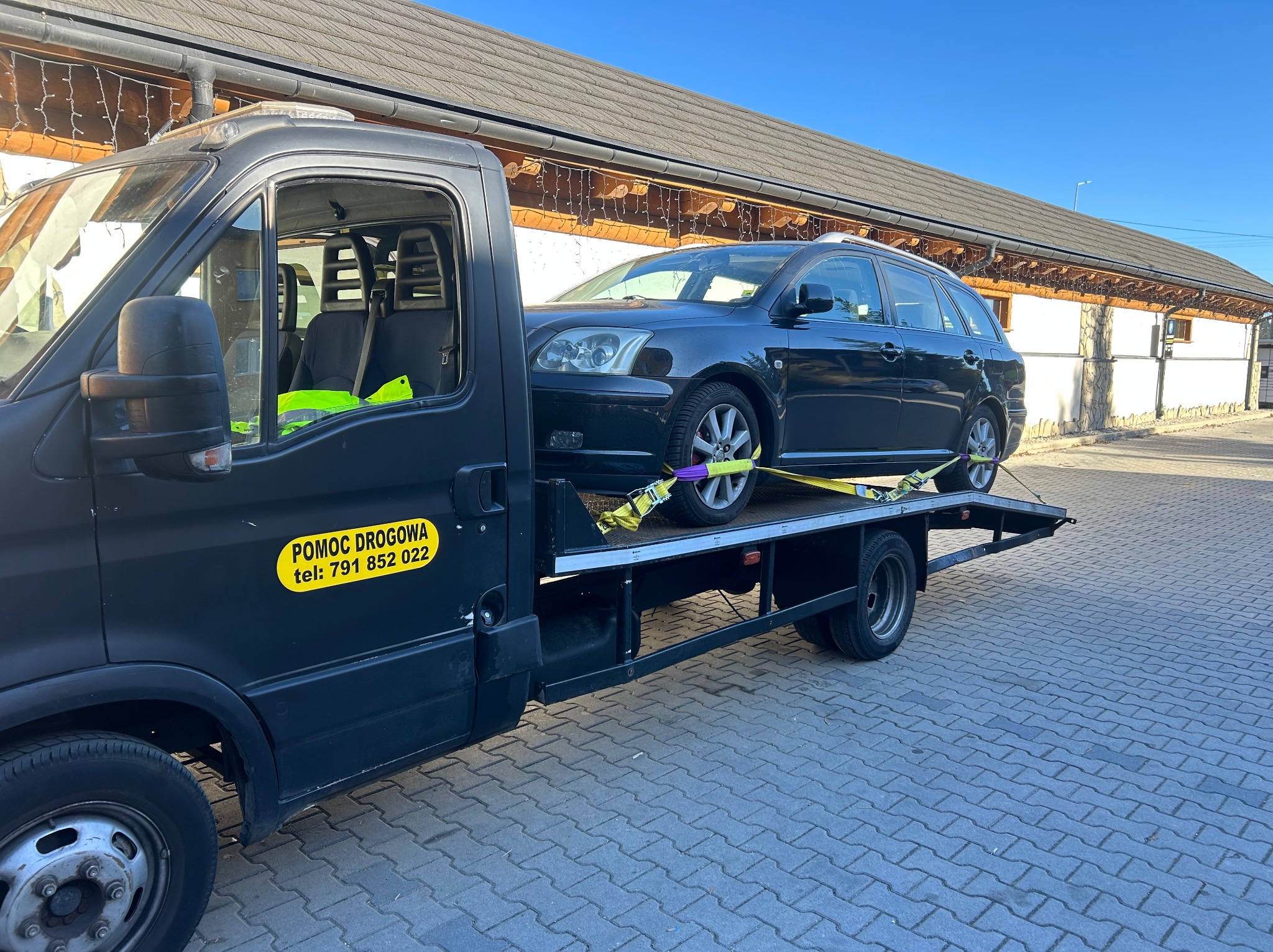 Czarny samochód osobowy kombi zabezpieczony pasami transportowymi na platformie pojazdu pomocy drogowej z żółtym napisem 'Pomoc Drogowa' i numerem telefonu, na tle budynku z dekoracjami świetlnymi.