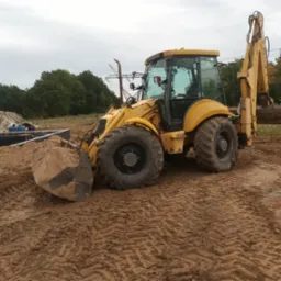 Żółta koparko-ładowarka LB 115 z łyżką pełną ziemi na placu budowy, widoczne fundamenty i zarys budynku w tle.