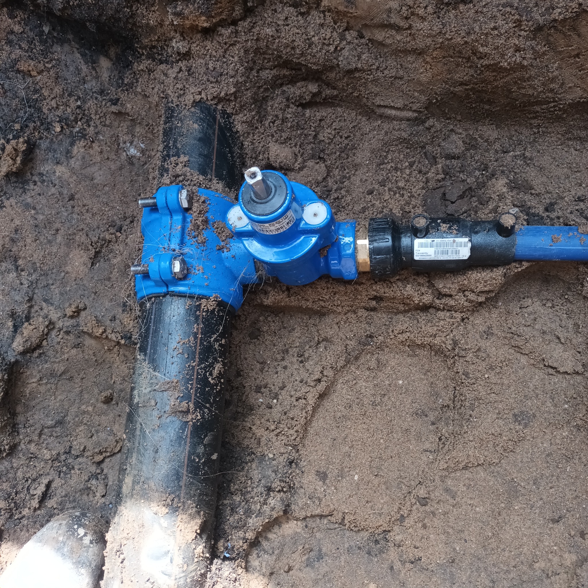 Niebieski zawór na rurze wodociągowej w wykopie ziemnym, widoczne ślady piasku i ziemi na elementach instalacji.