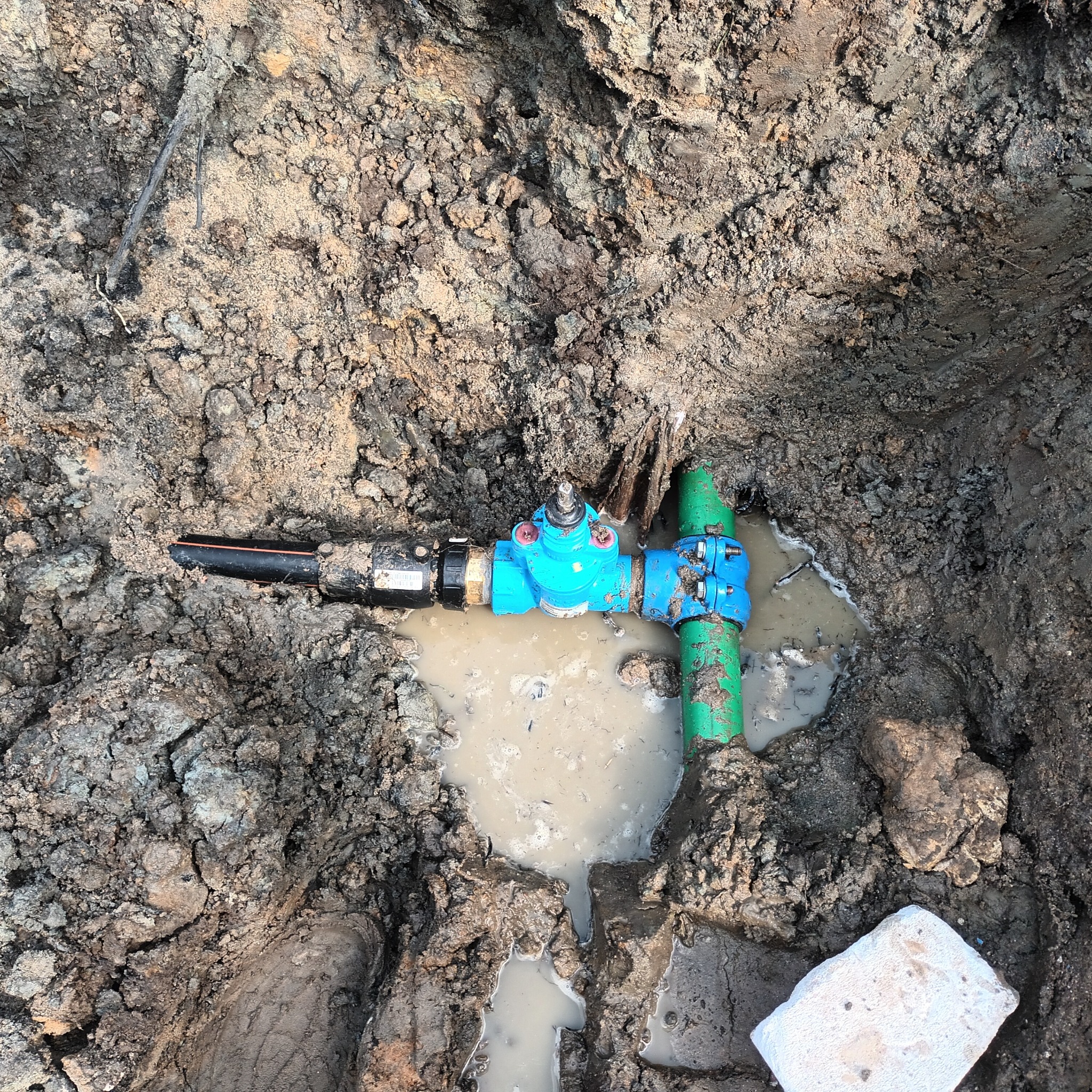 Fragment przyłącza wodno-kanalizacyjnego z niebieskim zaworem i zieloną rurą w wykopie wypełnionym błotnistą wodą, widoczne elementy instalacji zakopane w ziemi.