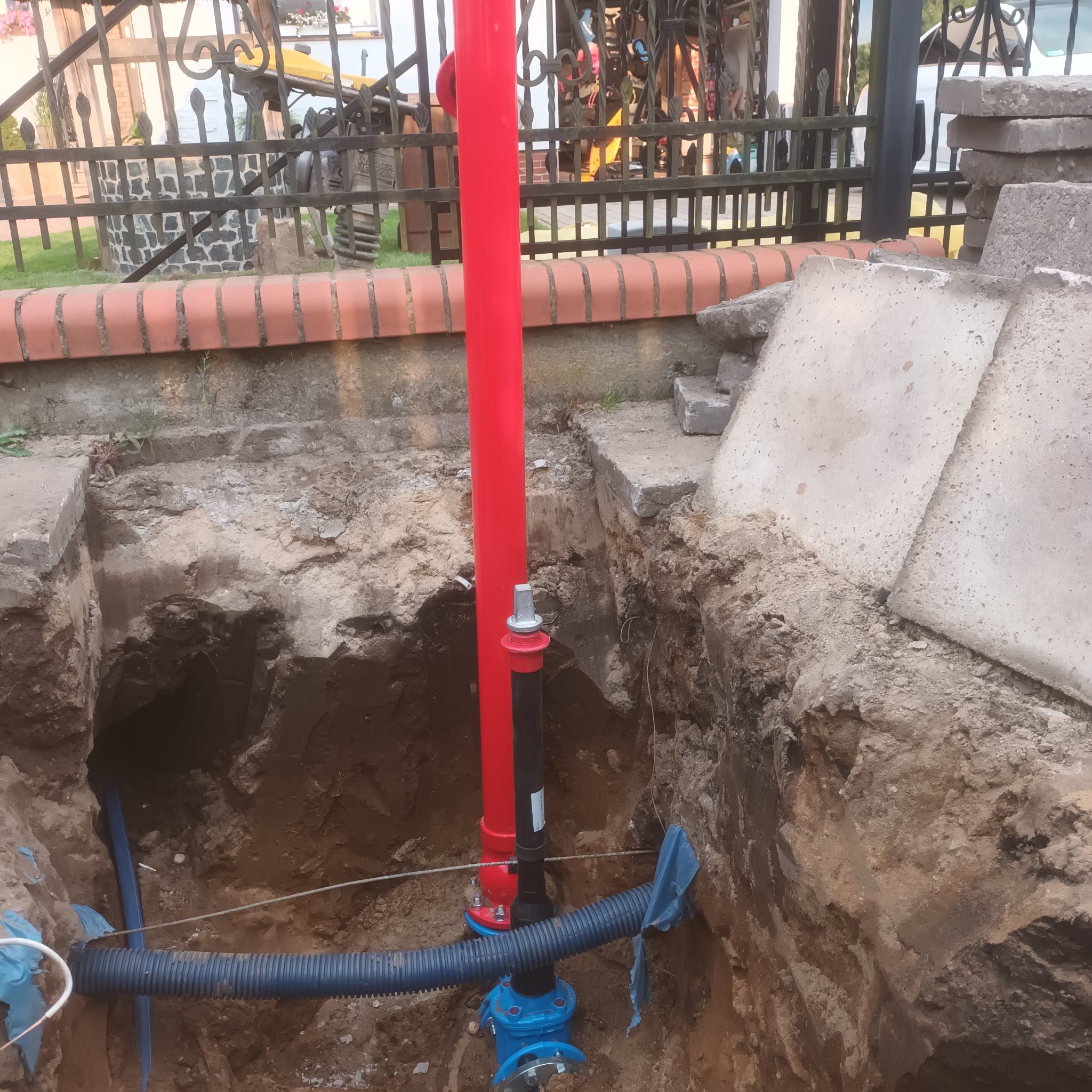Odkrywka ziemna z widoczną instalacją wodociągową: pionowa, czerwona rura połączona z niebieskim zaworem i karbowaną rurą drenażową, na tle ceglanego murku i kutego ogrodzenia.
