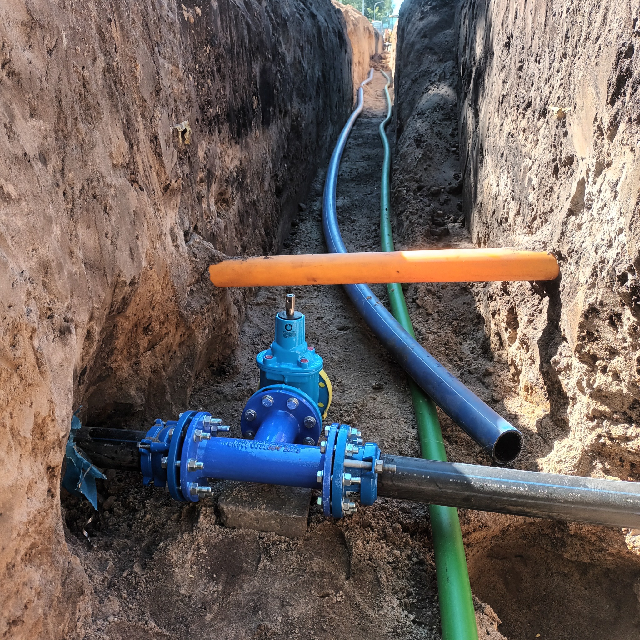 Wykop w ziemi z widocznymi rurami: niebieska z łącznikiem kołnierzowym, zielona oraz szara, dodatkowo widoczny zawór i pomarańczowa rura osłonowa.