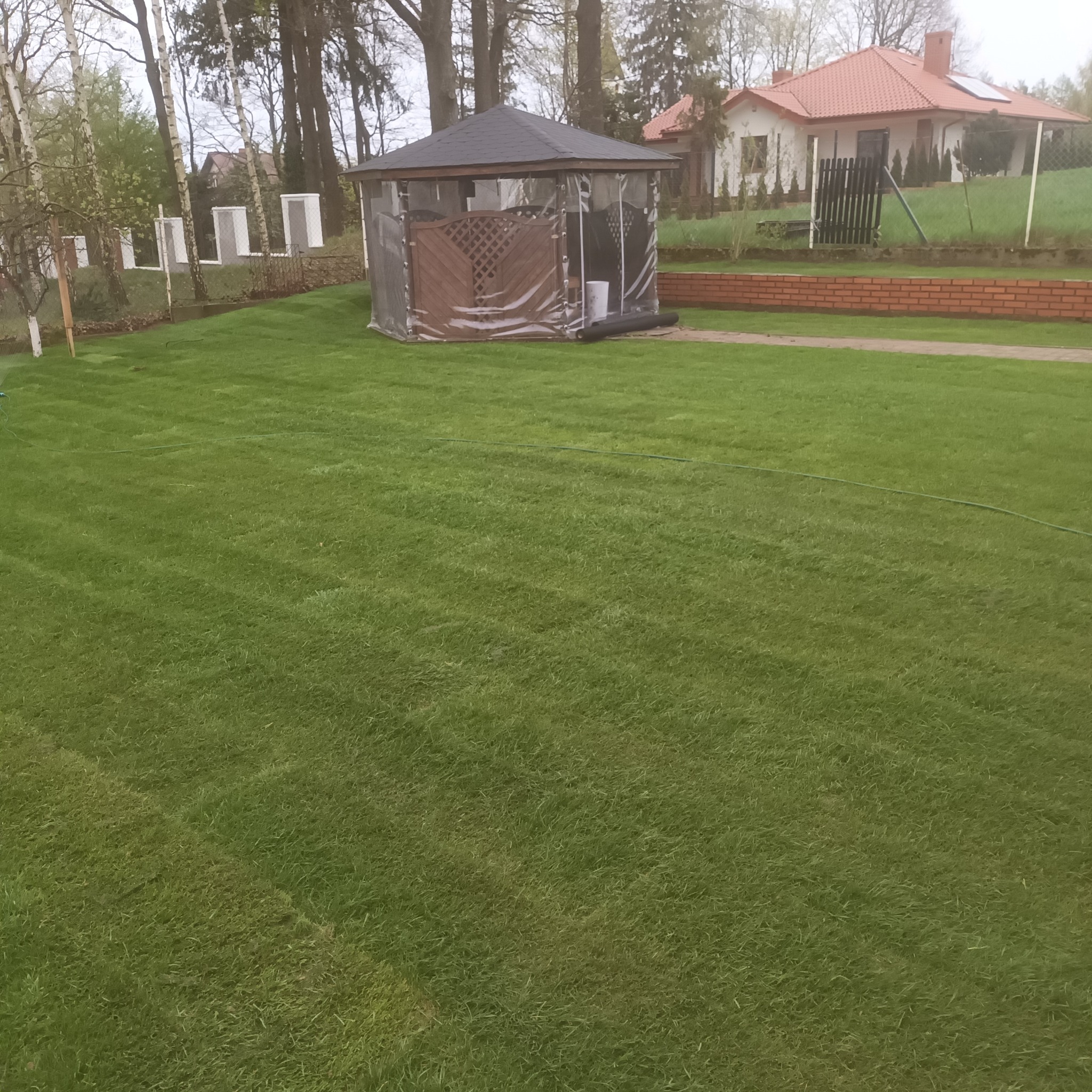 Starannie utrzymany trawnik z równo przystrzyżoną trawą, na którym stoi zadaszona altana z przezroczystymi ścianami, w tle widoczny ceglany murek i dom z czerwoną dachówką.