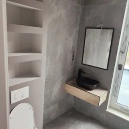 Nowoczesna łazienka z szarą okładziną imitującą marmur, wisząca czarna umywalka na drewnianej szafce, podwieszana toaleta i wnęka z półkami.