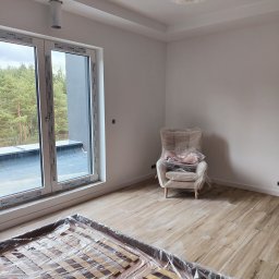 Wrzołart - Pokój w trakcie wykańczania: świeżo pomalowane białe ściany, podłoga imitująca drewno, okno z widokiem na las, meble zabezpieczone folią malarską.