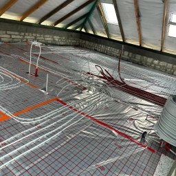 Wrzołart - Montaż ogrzewania podłogowego w poddaszu: białe i czerwone rurki ułożone na folii z czerwoną siatką, widoczne krokwie i okna dachowe.