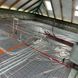 Montaż ogrzewania podłogowego w poddaszu: białe i czerwone rurki ułożone na folii z czerwoną siatką, widoczne krokwie i okna dachowe.