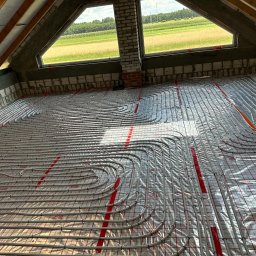 Wrzołart - Układanie rur ogrzewania podłogowego na folii z siatką w nowo budowanym poddaszu z widokiem na pole przez okno.